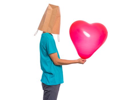 Valentines Day concept. Teen boy with paper bag over his head holds heart shaped balloon, isolated on white background. Boy holding symbol of love, family, hope. Teenager cover head with bag.