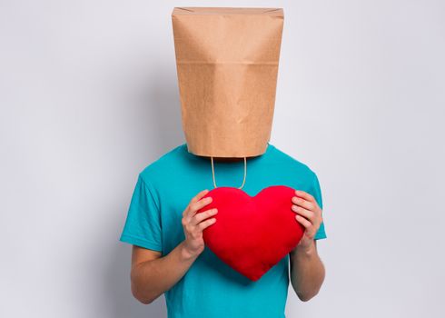 Valentines Day concept. Teen boy with paper bag over head holds red heart. Boy holding symbol of love, family, hope. Teenager cover head with bag posing in studio. Child pulling paper bag over head.