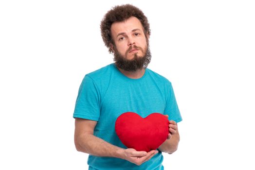 Happy Valentines Day. Crazy bearded Man with funny Curly Hair in blue T-shirt. Unhappy guy in Love, isolated on white background. Portrait of man with Red heart-shaped pillow, look at camera