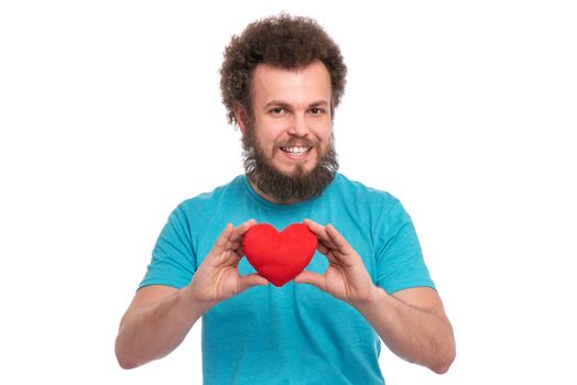 Happy Valentines Day. Crazy bearded Man with funny Curly Hair in blue T-shirt. Happy and silly guy in Love, isolated on white background. Portrait of Cheerful man with Red plush Heart, look at camera