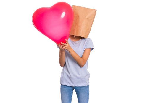 Valentines Day concept. Teen girl with paper bag over head holds red heart shaped balloon, isolated on white background. Child holding symbol of love, family, hope. Teenager cover head with bag.
