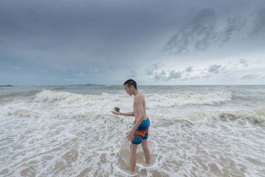 An Asian boy has to hold a stone on the beach with a cloudy sky.