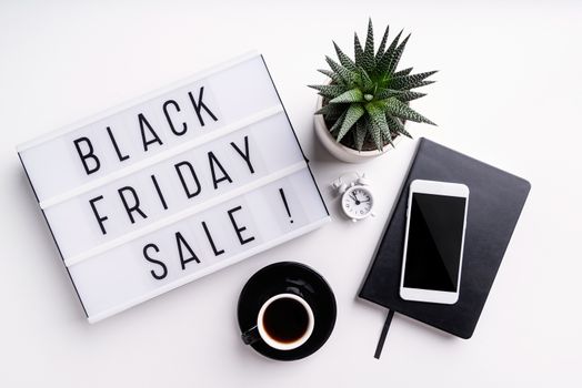 Black Friday shopping sale concept. Black Friday Sale words on lightbox with cup of coffee, mobile phone and potplant top view flat lay on white background