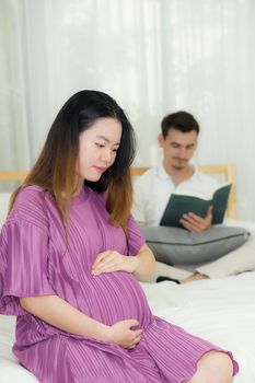 Asian pregnant people and expectation concept - happy pregnant woman sitting on bed and touching her belly at home with father reading book background.