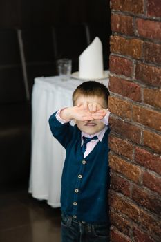 Blue-eyed boy in the room near the brick column covers his face with his hands