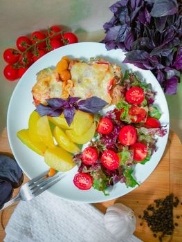 On a white plate there is a French style meat dish with slices of potatoes and cherry tomatoes and a green salad