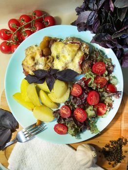 On a white plate there is a French style meat dish with slices of potatoes and cherry tomatoes and a green salad