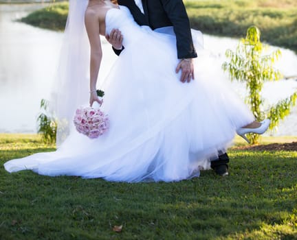 The groom leans his bride backwards for a kiss beside a lake in soft late afternoon light