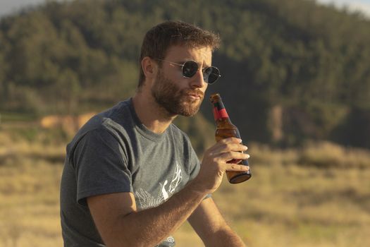 A young man, with glasses and a beard, drinks beer from a bottle, while enjoying the landscape and the sunset, in Galicia, northern Spain.