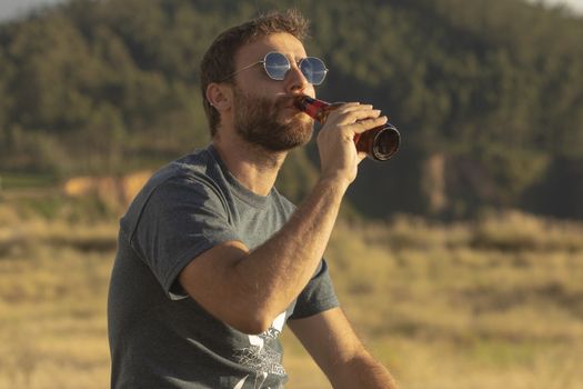 A young man, with glasses and a beard, drinks beer from a bottle, while enjoying the landscape and the sunset, in Galicia, northern Spain.