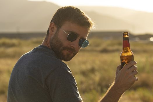 A young man, with glasses and a beard, drinks beer from a bottle, while enjoying the landscape and the sunset, in Galicia, northern Spain.