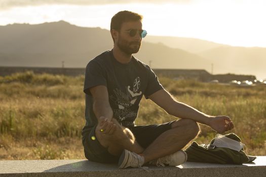 A young man, wearing glasses, meditates in a yoga posture, while enjoying the afternoon sun on a promenade in northern Spain.