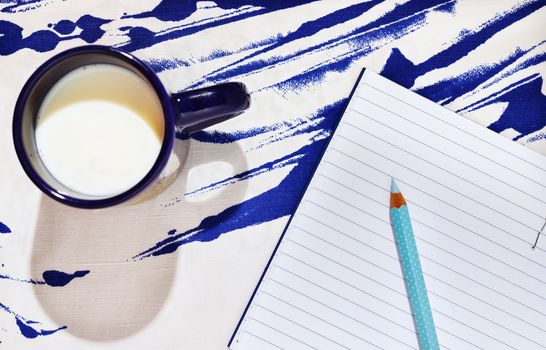 Blue mug with milk on blue-white cotton background , opened notebook with pencil in the background ,