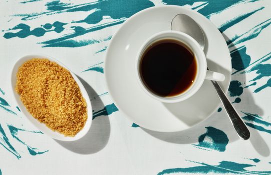 A white cup of coffee , white sugar bowl with brown sugar and spoon on colored cotton tablecloth