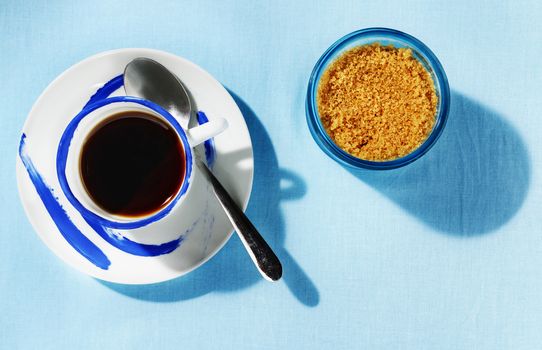 A cup of coffee with spoon and brown sugar in blue sugar bowl , blue tablecloth