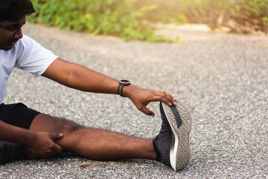 Close up Asian young athlete sport runner black man wear watch he sitting pull toe feet stretching legs and knee before running at outdoor street health park, healthy exercise before workout concept