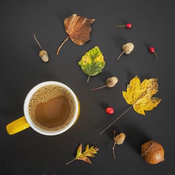 Cup of coffee surrounded by autumn leaves, holly, and acorns on a dark grey background