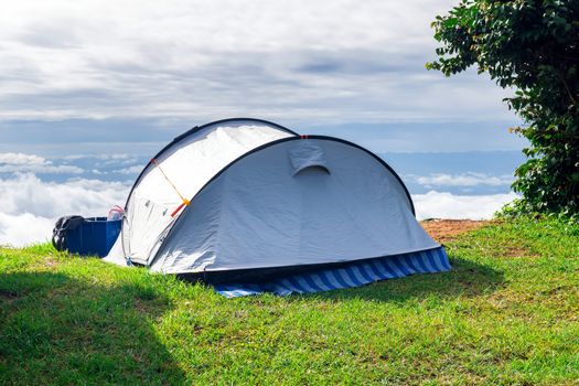 Closeup a camping tent that is set up on a grass field at the highest point of a high mountain with a backdrop of sky and clouds.