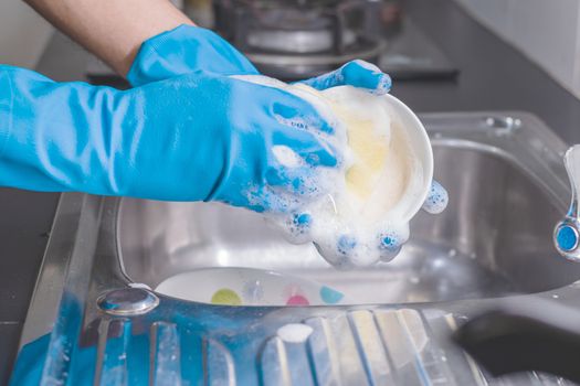 Close up of a man wearing a blue rubber glove was washing the cup with a dish washing liquid in the sink in the kitchen.