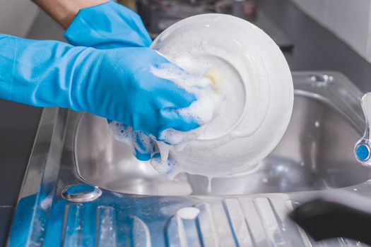 Close up of a man wearing a blue rubber glove was washing the cup with a dish washing liquid in the sink in the kitchen.