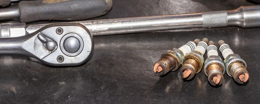 Shot of used spark plugs on a table with socket wrench blurred in the background. Another tool lying next to them on the table. Car repair shop.