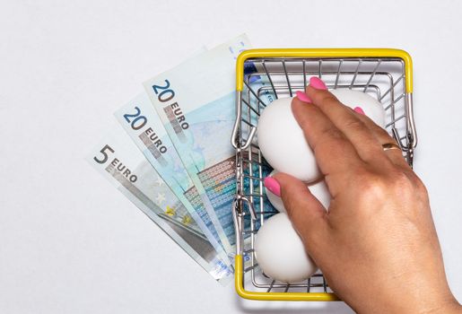 Fresh eggs in a shopping cart with various euro bills underneath it and womans hand reaching for eggs. Shopping, purchasing, and food delivery concept. White background. Close up shot. Isolated.