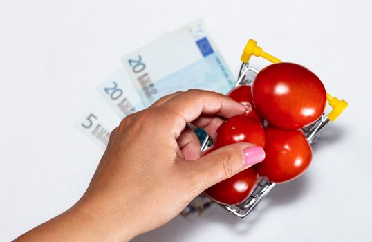 Shot of tomatoes in shopping cart isolated on white background with euro bills under it and womans hand reaching for tomato. Top view. Ripe tasty red tomatos in shopping cart. Tomato trading concept. Online shopping concept.