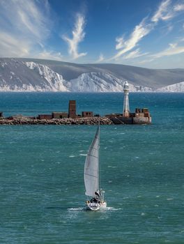 High angle shot of a white sailboat sailing in Weymouth Bay, UK. Coast line with white cliffs and gorgeous cloudy blue sky in the background. Lighthouse at the harbour entrance. Sport and recreation concept. Vertical, portrai format.