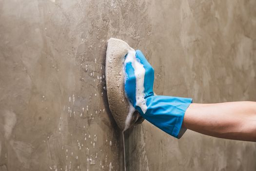 Closeup hand wearing blue rubber gloves is using a sponge cleaning on the concrete wall.