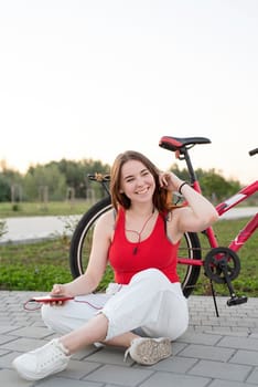 Laughing teenager girl sitting next to her bike in the park listening to the music