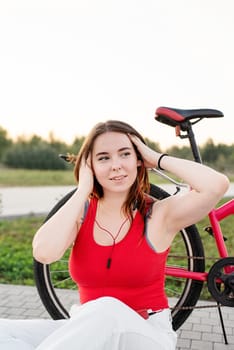Teenage girl sitting next to her bike listening to the music in the park at sunset