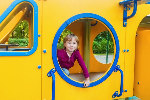 The girl looks out from the circle of the slide on the playground looks at the camera and smiles.