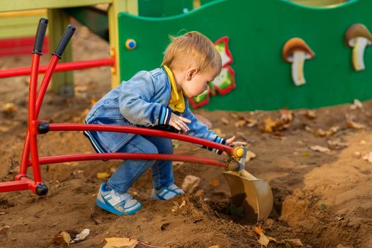 Little boy in denim clothes plays with a toy excavator on the playground in the autumn park.