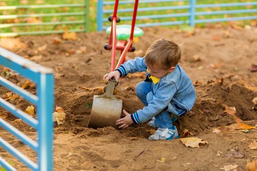 Little boy in denim clothes plays with a toy excavator on the playground in the autumn park.