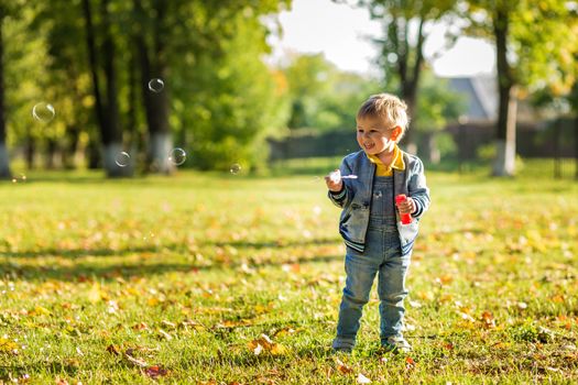 A little boy in denim clothes blows bubbles in the autumn park.