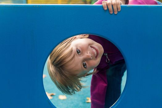 The girl looks out from the circle of the slide on the playground looks at the camera and smiles.