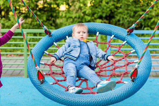 .The sister is swinging her brother on the swing in the playground. The girl has a broken arm.