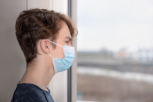Concept of coronavirus quarantine. Child wearing medical protective face mask during flu virus, looking out of window. COVID-19 - self isolation. Teen boy forced to stay at home.