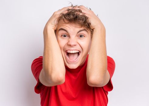 Portrait of surprised teen boy, on grey background. Funny child looking at camera in shock or amazement touching his head with hands. Handsome caucasian young teenager.