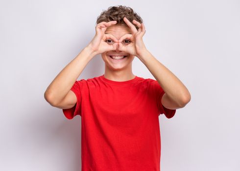 Portrait of handsome teen boy making Ok Gesture with hand on eyes looking through fingers. Happy smiling child showing glasses okay.