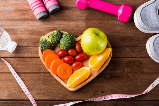 Top view of fresh fruits and vegetables in heart plate wood (apple, carrot, tomato, orange, broccoli) and dumbbells, sport shoes sports equipment on wooden table, Healthy lifestyle diet food concept