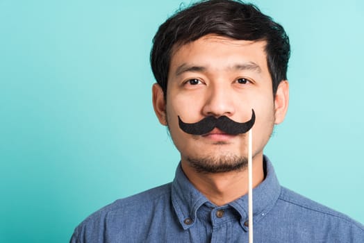 Portrait Asian happy handsome man posing he holding a funny mustache card or vintage fake moustaches on his mouth, studio shot isolated on blue background, Fathers day, November day concept