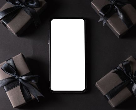 Black Friday sale shopping concept, Top view of gift box wrapped black paper and black bow ribbon present around smartphone blank screen, studio shot on dark background