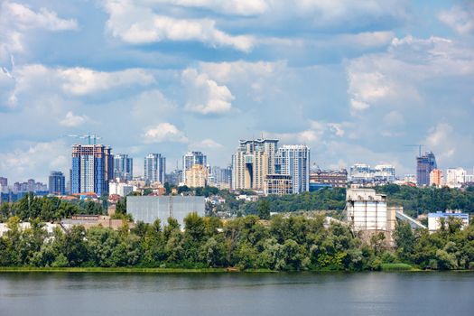 The cityscape of the right bank of Kyiv with the Dnipro River and an industrial complex on the foreground and a view of a new residential area with high-rise buildings on the horizon on a warm summer day.