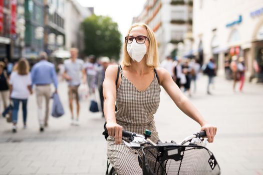 Woman riding bicycle on city street wearing medical face mask in public to prevent spreading of corona virus. New normal during covid epidemic. Social responsibility.