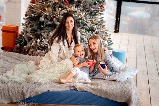 Happy cheerful family near Christmas tree. Mother and kids wearing pajama having fun near tree in the morning. Merry Christmas and Happy Holidays concept