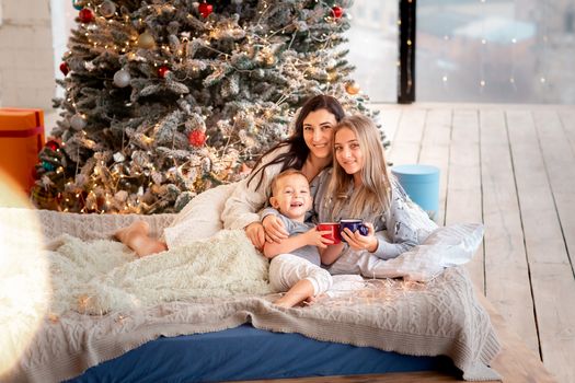 Happy cheerful family near Christmas tree. Mother and kids wearing pajama having fun near tree in the morning. Merry Christmas and Happy Holidays concept