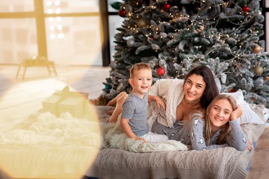 Happy cheerful family near Christmas tree. Mother and kids wearing pajama having fun near tree in the morning. Merry Christmas and Happy Holidays concept