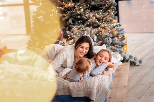 Happy cheerful family near Christmas tree. Mother and kids wearing pajama having fun near tree in the morning. Merry Christmas and Happy Holidays concept