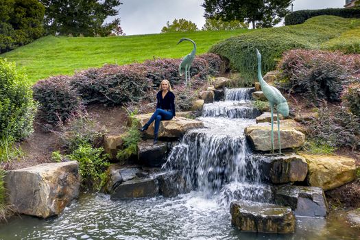 A gorgeous blonde model sits near a waterfall at a local park